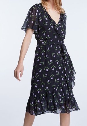 Kvinde iført en sort slå-om kjole med lilla tulipanprint, korte flagrende ærmer og flæsekant, bundet i taljen med et stofbælte.