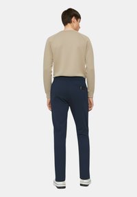 Camicia beige a maniche lunghe con maniche raglan, abbinata a pantaloni blu navy con tasca sul retro e vita elastica, indossata con sneakers bianche.