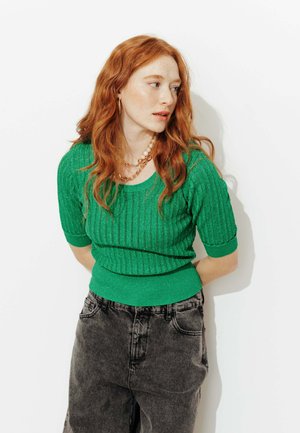 Femme aux cheveux roux portant un pull vert côtelé à manches courtes, des colliers superposés en or et perles, et un jean noir délavé taille haute.