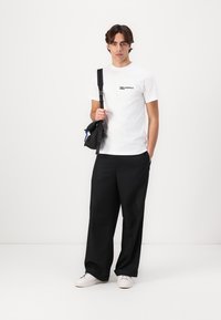 Weißes T-Shirt mit kurzen Ärmeln und schwarzem Logo, kombiniert mit lockeren schwarzen Hosen. Schwarze Umhängetasche, weiße Sneaker. Schlichter Hintergrund.