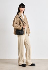 Trench coat beige con bottoni neri, abbinato a pantaloni larghi color crema. Borsa a tracolla nera con un logo evidente. Scarpe nere con plateau.