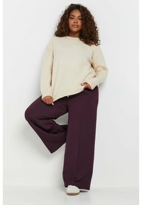 Beiges, übergroßes Sweatshirt mit weichem Material und rundem Ausschnitt. Kombiniert mit weiten lila Hosen. Die Schuhe sind weiß mit einer braunen Sohle.