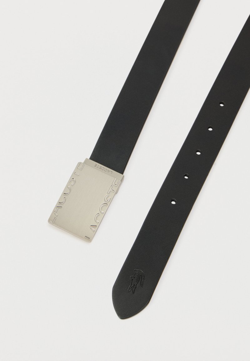 Ceinture en cuir noir avec une boucle rectangulaire en métal argenté embossée du mot "LACOSTE", équipée de trous pour réglage et d'un détail logo.