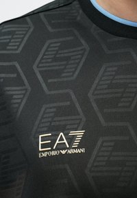 Melnā sporta krekls ar reljefu sešstūra rakstu, zilu apkakles detaļu un zelta EA7 Emporio Armani logo krūšu rajonā.