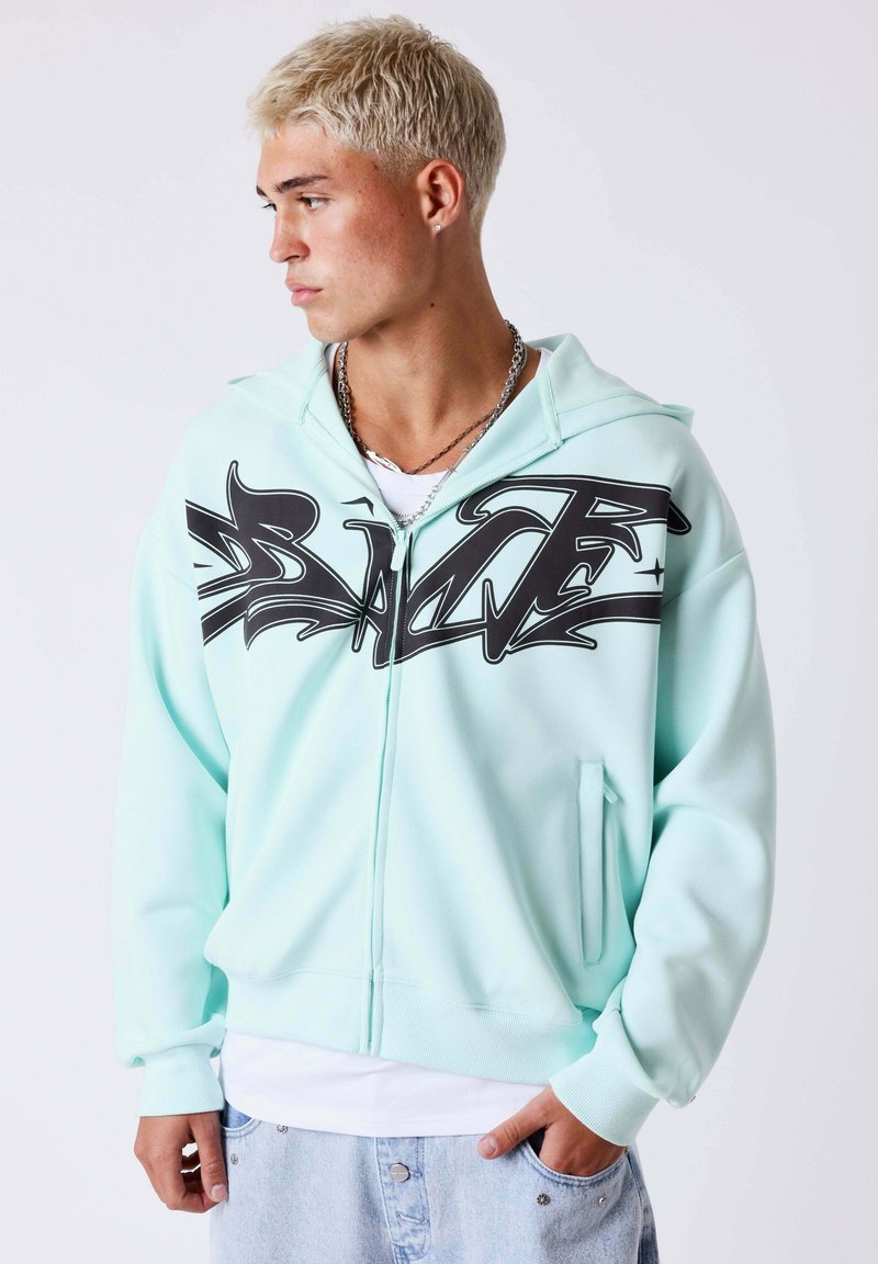 Jeune homme aux cheveux courts décolorés portant un sweat à capuche zippé bleu clair avec un imprimé graphique noir, une chemise blanche et un jean en denim clair.