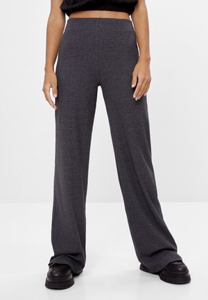 Person trägt hochgeschnittene, weit geschnittene dunkelgraue Hose und schwarze Plateauschuhe, steht auf weißem Boden mit einfarbig weißem Hintergrund.