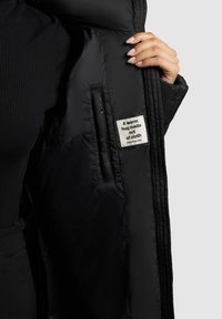 Fekete puffjaket, steppelt textúrával, belső címkével, amely szöveget tartalmaz. Közelkép egy kézről, ami az nyitott jakettre fog.
