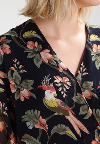 Blond kvinna som bär en svart blus med färgglada blomm- och papegoja-mönster, i röd, gul och grön nyans.