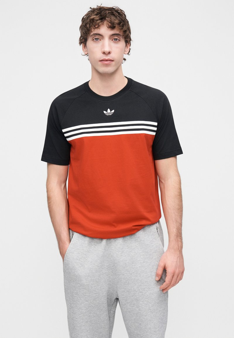 T-shirt homme à col rond avec un haut noir et un bas rouge, orné de trois bandes horizontales blanches. Confectionné en tissu lisse.