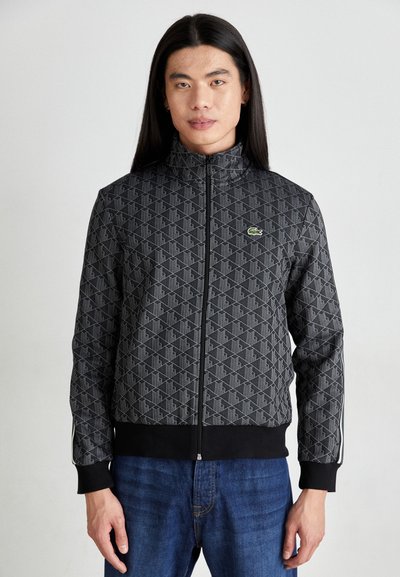 Veste noire à fermeture éclair avec motif géométrique, poignets et bord-côte côtelés, arborant un petit logo vert sur la poitrine. Portée avec un jean bleu.