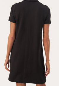 Donna che indossa un abito nero a maniche corte con colletto, vista da dietro su uno sfondo chiaro e uniforme.