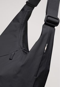 Borsa a tracolla nera realizzata in tessuto morbido e resistente all'acqua, con una tasca con zip, tracolla regolabile e un piccolo logo decorativo.