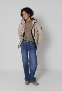 En beige pufferjacka med svart dragkedja, grå topp, blå vida jeans och beige skor, stående mot en enhetlig bakgrund.