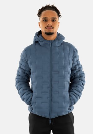 Giacca blu imbottita con cappuccio, caratterizzata da un motivo superficiale testurizzato, chiusura frontale con zip e polsini elasticizzati. Indossata sopra pantaloni neri.