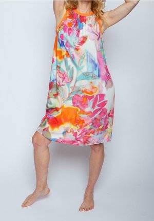 Vrouw draagt een kleurrijke mouwloze knie-lange jurk met abstracte bloemenpatronen, staat blootsvoets met één been opgeheven.