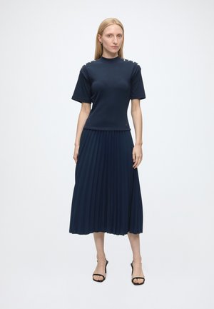 Vrouw staat en draagt een marineblauw shirt met korte mouwen met knopen op de schouders en een bijpassende geplooide midi-rok met zwarte sandalen met hak.