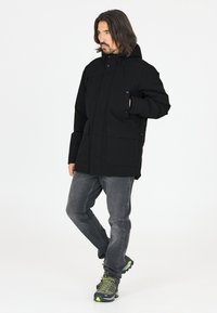 Sort parka-jakke med hætte, lynlås-lukning og to store forlommer. Båret med grå jeans og sorte og grønne vandresko.