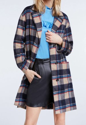 Frau mit einem beigen, marineblauen und roten karierten Mantel über einem blauen Rüschenshirt und einem schwarzen Lederrock, mit einer Hand in der Tasche und der anderen Hand, die den Mantel hält.