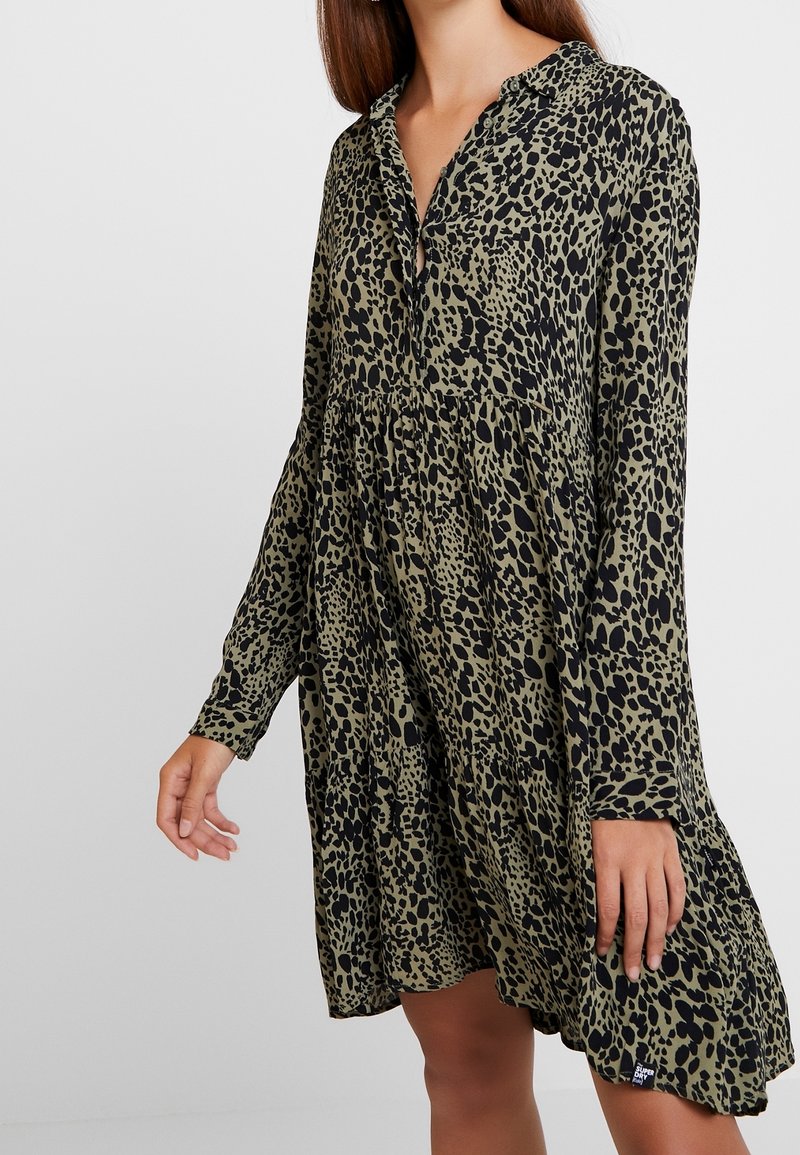 Šaty s dlouhým rukávem v olivově zelené barvě s černým leopardím potiskem. Mají zapínání na knoflíky u výstřihu a volný, vrstvený střih. Měkká textura.