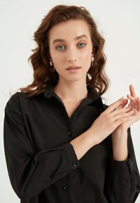 Camicetta nera con colletto, realizzata in un tessuto liscio. Il modello ha capelli ondulati e indossa orecchini a cerchio, con le mani posizionate in modo elegante.