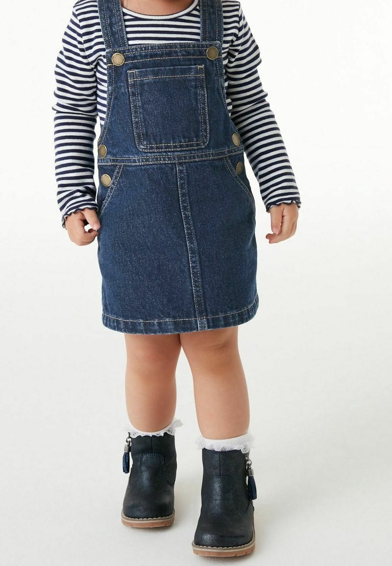 Denim overall dress dengan kantong, tombol kuningan, dan hem yang melebar. Dipadukan dengan kemeja lengan panjang bergaris navy dan sepatu bot pergelangan kaki hitam.