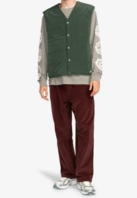 Gevoerde groene vest met een V-hals ontwerp, met drukknoopsluitingen en een gestructureerd oppervlak, gedragen over een grijze lange mouwen shirt en maroon broek.