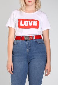 Femme portant un t-shirt blanc avec «LOVE» en lettres rouges, un jean bleu taille haute et une ceinture rouge avec une boucle métallique.