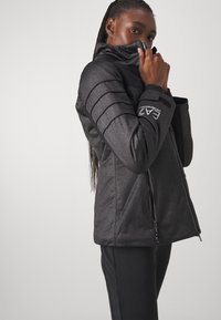 Mustapintainen tekoturkisjakku, jossa on seisomakaulus, vetoketjukiinnitys ja ribbattuja olkaimia. Sisältää logon hihassa ja elegantin muotoilun.