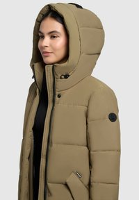 Giacca puffer kaki con grande cappuccio, zip frontale e bottoni a pressione. Presenta una patch con logo sulla manica e materiale testurizzato.