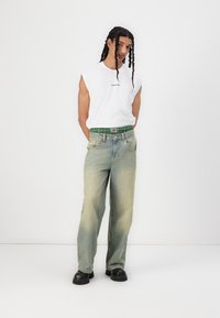 Hvit ermeløs skjorte med logo, lyseblå baggy jeans med grønn mønstrete midjebånd, og svarte klumpete sko. Enkel og casual design.