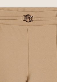 Pantaloni beige in tessuto texturizzato, con design frontale piatto e una fibbia circolare con logo in rilievo e pietre decorative.