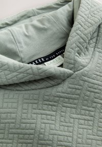 Vêtement en tissu vert clair texturé avec un gros plan sur le col et des étiquettes de la marque Ted Baker visibles à l’intérieur.