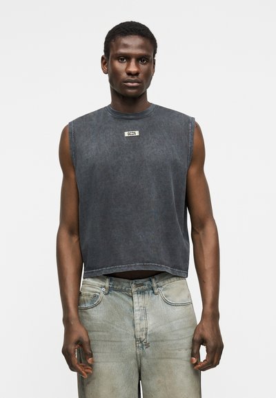 Νεαρός άνδρας που φοράει ένα ξεβαμμένο μαύρο αμάνικο crop top με το λογότυπο "GCDS" και ανοιχτόχρωμο μπλε ξεθωριασμένο τζιν, στέκεται μπροστά σε ένα απλό φόντο.