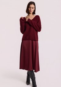 Burgundowy, oversized sweter z dekoltem w serek i długimi rękawami, w połączeniu z burgundową satynową spódnicą midi oraz czarnymi kozakami do kolan.