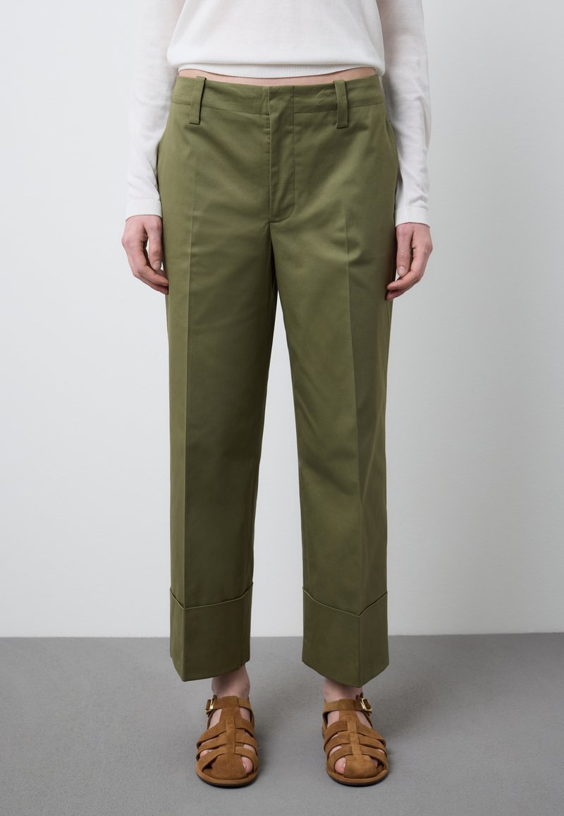 Person trägt olivgrüne, gerade geschnittene Hosen mit breiten Umschlägen, ein weißes Langarmshirt und braune Riemchensandalen und steht auf einem grauen Boden.