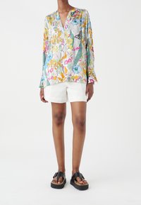 Personne portant un chemisier à manches longues floral coloré, un short blanc et des sandales à plateforme noires, debout devant un fond blanc uni.