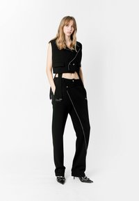 Chaqueta negra corta con diseño asimétrico y bolsillos frontales, combinada con pantalones negros ajustados que tienen una cremallera diagonal y bolsillos laterales.