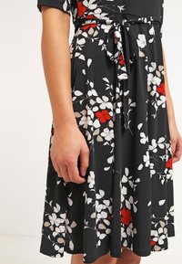 Gros plan d'une personne portant une robe noire avec des motifs floraux blancs et rouges, avec une ceinture nouée à la taille.