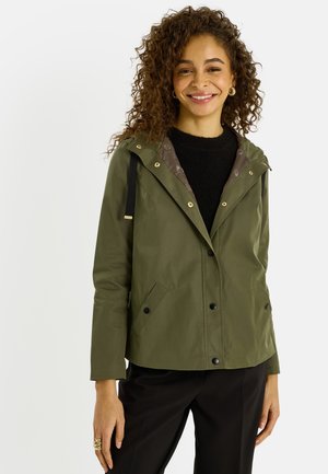 Femme souriante aux cheveux bouclés portant des boucles d'oreilles créoles en or, une veste vert olive sur un pull noir, et un pantalon noir sur un fond clair.