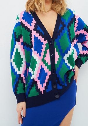 Femme portant un cardigan coloré à motif géométrique avec de gros boutons sur une jupe asymétrique bleue.