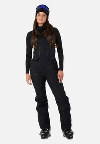 Czarny kombinezon narciarski z regulowanymi ramiączkami, licznymi kieszeniami na zamek i wzmocnionymi kolanami, w połączeniu z czarnymi śniegowcami i pasującą czapką.