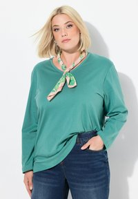 Langärmliges, türkises Baumwoll-T-Shirt mit lässiger Passform, kombiniert mit dunklen Jeans. Ein floral gemusterter, pinkfarbener Schal, der am Hals gebunden ist, sorgt für das gewisse Etwas.