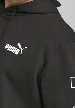 Gros plan sur une personne portant un sweat à capuche noir avec un logo Puma blanc sur la poitrine et un texte blanc supplémentaire sur la manche.