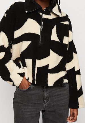 Mujer con sudadera polar de patrón abstracto en blanco y negro con cremallera de medio cierre, con la mano en el bolsillo de unos vaqueros gris oscuro, sobre un fondo liso.