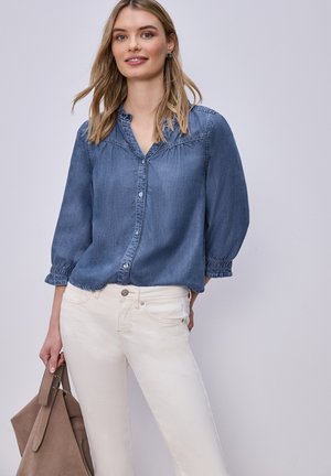 Femme portant un chemisier en denim bleu avec des boutons et un pantalon blanc, tenant un sac à main en daim marron sur un fond clair uni.