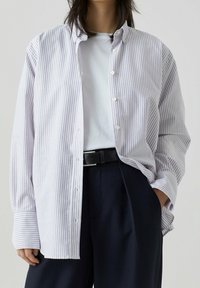 Camicia bianca con righe viola, colletto button-down, vestibilità rilassata, maniche lunghe, abbinata a una t-shirt bianca e pantaloni scuri, cintura nera.