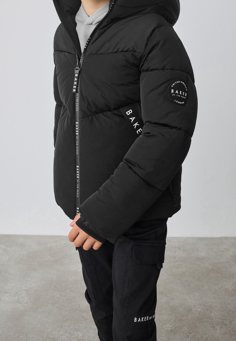 Enfant portant une doudoune noire et un pantalon noir, tous deux de la marque "Baker", debout à l’intérieur sur un sol gris avec un fond clair uni.