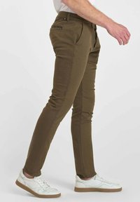 Zijaanzicht van een persoon die in slim-fit olijfgroene broeken en witte sneakers met bruine zolen loopt tegen een effen achtergrond.