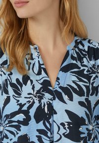 Blusa con stampa floreale caratterizzata da uno sfondo blu e grandi fiori neri. Ha un collo a V e una texture morbida con leggere arricciature sulle spalle.