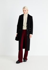 Zwarte lange jas met ingesneden revers en twee knopen, gecombineerd met een crèmekleurige coltrui en bordeauxrode wijde broeken. Getextureerde zwarte tas en chunky schoenen.